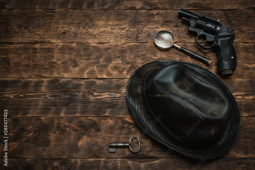 Detective or spy agent wooden table flat lay background with copy space. Handgun, black leather hat, magnifying glass and a rusty key on the work table. Detective or spy agent wooden table flat lay background with copy space. Handgun, black leather hat, magnifying glass and a rusty key on the work table.