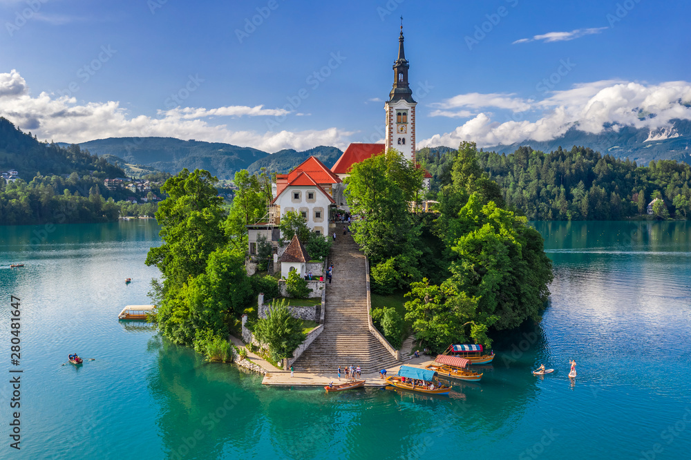 Bled, Slovenia - Aerial view of beautiful Pilgrimage Church of the Assumption of Maria on a small island at Lake Bled (Blejsko Jezero) and lots of Pletna boats on the lake at summer time with blue sky - obrazy, fototapety, plakaty 