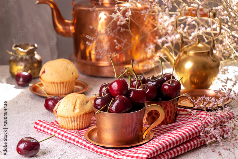 Sweet cherries in cooper cups and muffins against of background with old cooper teapot. Summer dessert.