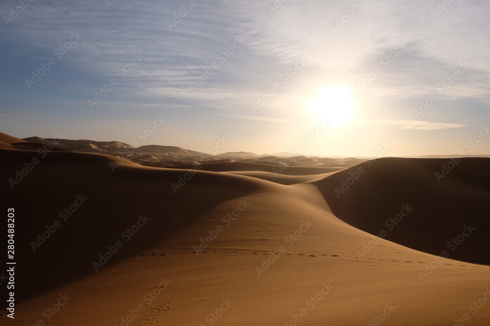 wild brown Sahara desert sand dunes at sunset. Strong shining sun with white cloud. footsteps and shadows. Saharan, sandy near Merzouga in Morocco