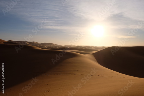wild brown Sahara desert sand dunes at sunset. Strong shining sun with white cloud. footsteps and shadows. Saharan  sandy near Merzouga in Morocco