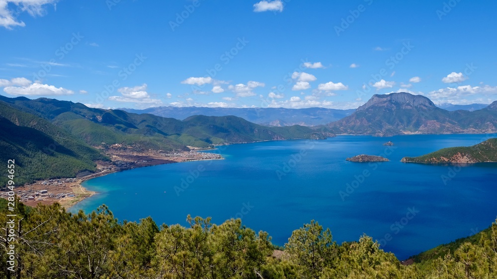 deep blue lake surrounded by green mountains. Green trees foreground. Sunny blue sky with white clouds. high angle view of Lugu lake in Yunnan China