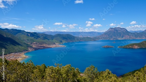 deep blue lake surrounded by green mountains. Green trees foreground. Sunny blue sky with white clouds. high angle view of Lugu lake in Yunnan China © Robert