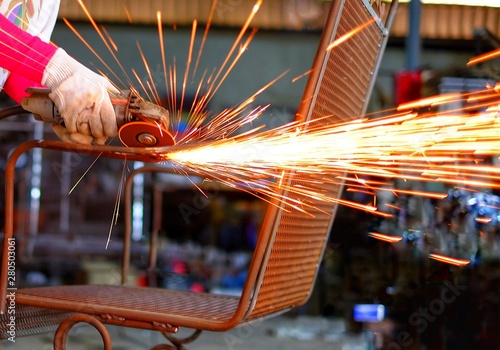 A Indian woman wearing a pink long sleeve jacket and protective gloves is welding and grinding a furniture chair with sparks flying