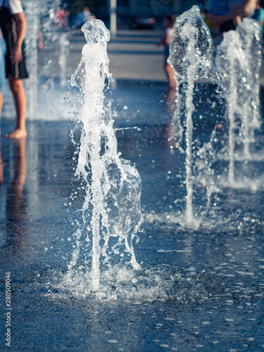 Water streams of dry fountain on city square, close up view. Kids cooling among water sprays. Summer cityscape background. Selective soft focus. Blurred background