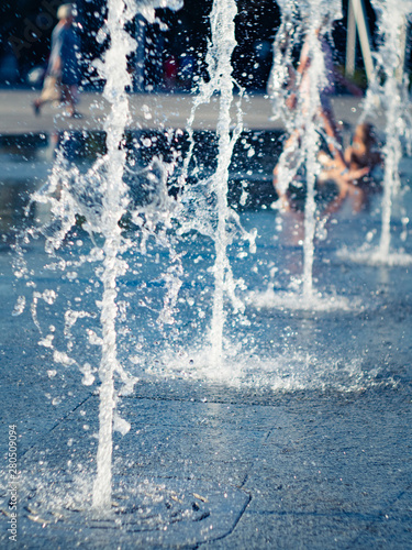 Water drops from dry fountain splashing on granite square coating, close up view. Children cooling in water sprays. Summer cityscape. Selective soft focus. Blurred background