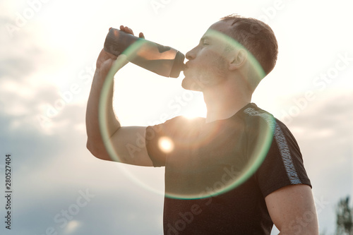 portrait of a young Caucasian guy in a black t-shirt and black shorts drinking water from a bottle after or before training
