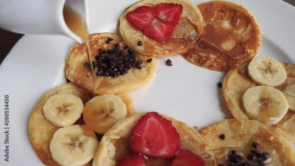 Pouring Maple Syrup in a circular motion over an elegant arrangement of pancakes with sliced strawberries, banana, and chocolate. A stabilized 60fps 4k close up shot.