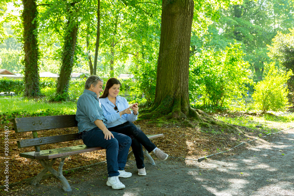 Lady shows some content of her phone to a man when sitting on a bench in a city park Lady shows some content of her phone to a man when sitting on a bench in a city park