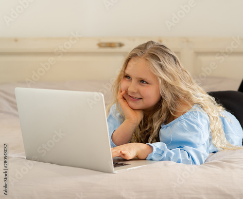 Beautiful cute little blonde girl playing and surfing the internet on laptop at home.