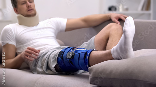 Man in in foam cervical collar and neoprene knee brace lying on sofa, pain