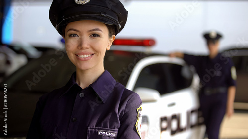 Canvas Smiling asian policewoman posing to camera against partner near patrol car, duty