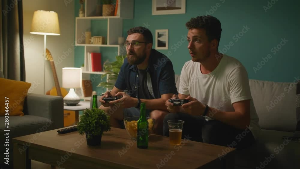 In the Living Room Two Friends Sitting on a Couch Playing Competitive Video Game In the Living Room Two Friends Sitting on a Couch Playing Competitive Video Game