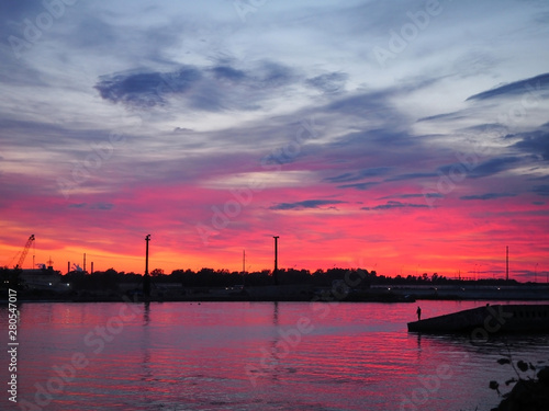 A beautiful red and yellow sunset over the city is reflected in the water of the river with the .silhouette of a fisherman in the background. Panoramic landscape on the dusk.