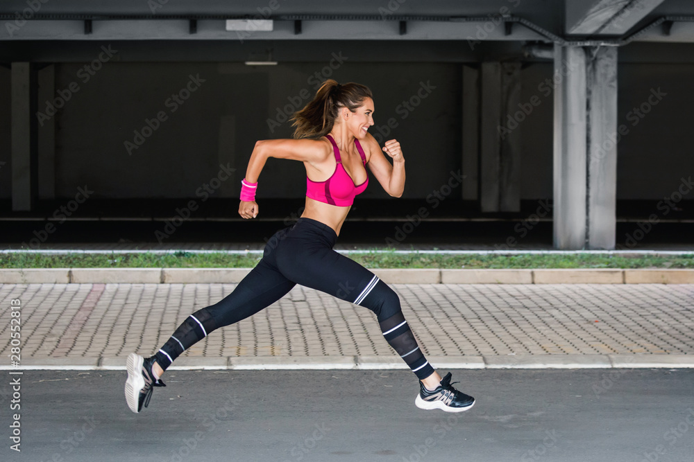 Young woman with fit body jumping and running against grey background. Female model in sportswear exercising outdoors.