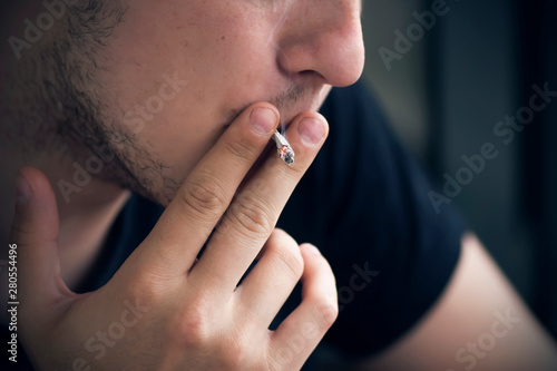 Wallpaper Mural A guy in dark clothes with a stubble on his face is inhaling nicotine from a burning cigarette, holding it with two fingers. Legal harm to health. Torontodigital.ca