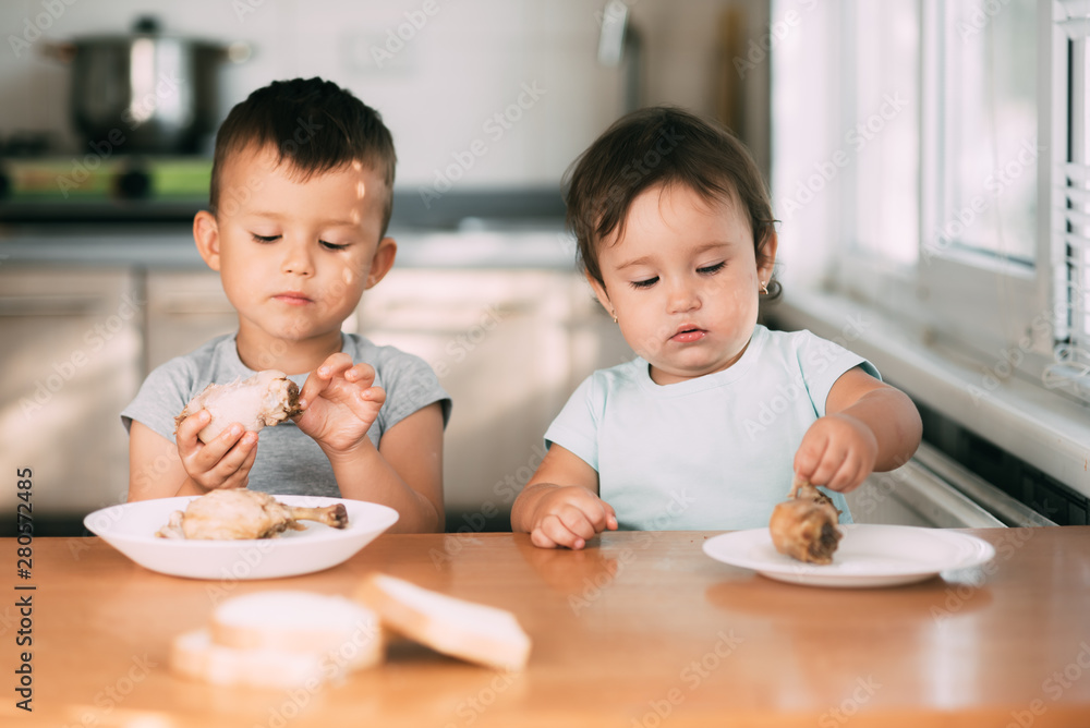 Kids boy and girl , brother and sister in the kitchen having fun and eating chicken with appetite Kids boy and girl , brother and sister in the kitchen having fun and eating chicken with appetite