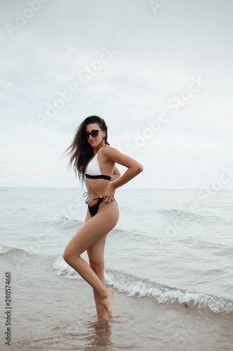Amazing girl in swimsuit with a beautiful sports body walking and posing on a white sand beach. A tanned young woman with curly hair is resting and sunbathing. Charming model posing near the sea. © Tetiana Moish
