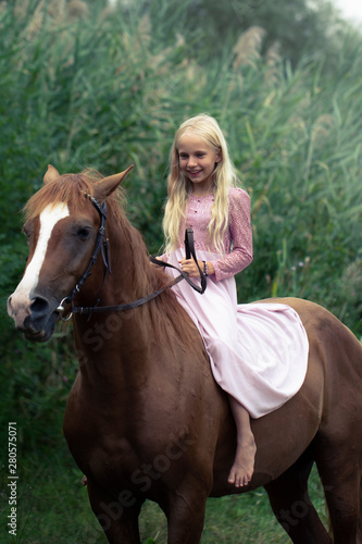 beautiful smiling caucasian girl with blonde hair in a pink dress rides a brown horse in the forest. natural beauty concept. happy childhood poster