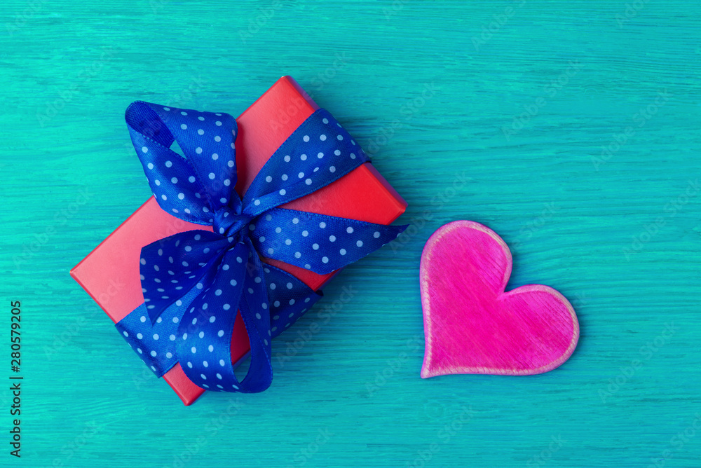 Obraz premium Red gift box with wide blue spotted ribbon and pink heart on turquois wooden background