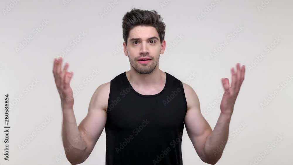 Displeased young bearded sporty man in black shirt becoming disappointed and gesturing with hands while looking at the camera over gray background isolated Displeased young bearded sporty man in black shirt becoming disappointed and gesturing with hands while looking at the camera over gray background isolated
