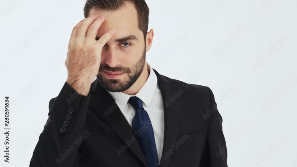 Close up view of handsome young businessman in black suit and blue tie smiling and fixing his hair while looking flirty at the camera over gray background isolated Close up view of handsome young businessman in black suit and blue tie smiling and fixing his hair while looking flirty at the camera over gray background isolated