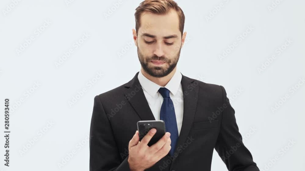 Serious young businessman in black suit and blue tie taking his smartphone out of the pocket and becoming angry and disappointed while having a call over gray background isolated Serious young businessman in black suit and blue tie taking his smartphone out of the pocket and becoming angry and disappointed while having a call over gray background isolated