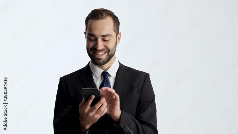 Happy young businessman in black suit and blue tie smiling while using smartphone over gray background isolated Happy young businessman in black suit and blue tie smiling while using smartphone over gray background isolated