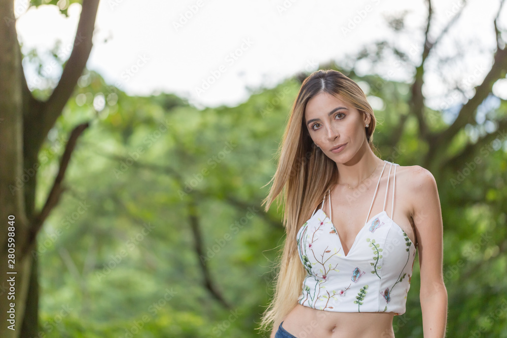 Lady model poses for portrait outdoors with nature at the background. She is wearing a casual top and blue jeans. She is fit. She has long smooth blonde hair, big smile and brown clear eyes.