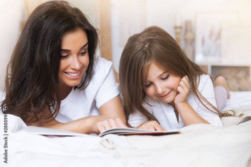 Happy family - close-up portrait of smiling mother and daughter reading in bed early in the morning in the white Scandinavian-style interior Happy family - close-up portrait of smiling mother and daughter reading in bed early in the morning in the white Scandinavian-style interior