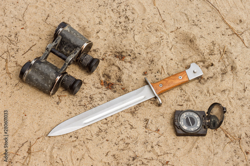 Fotografija Knife bayonet with wooden handle lies on the sand with old military binoculars and old compass