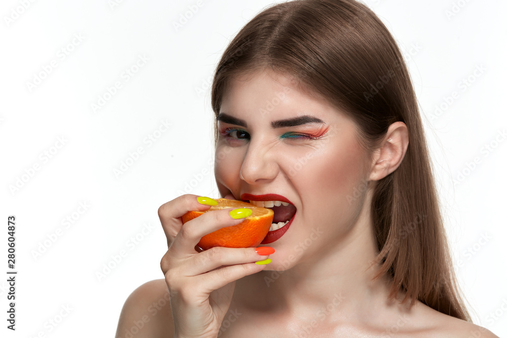 Closeup portrait of a beautiful young woman with bright color make-up biting half of an orange near the face. Closeup portrait of a beautiful young woman with bright color make-up biting half of an orange near the face.