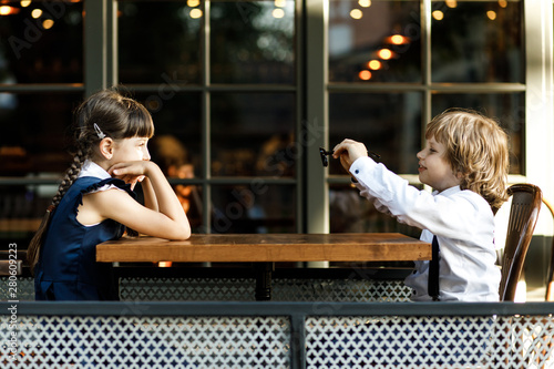 Cute blond guy and pretty girl, school years, dressed in school uniform, spends her time in the summer cafe on the street.