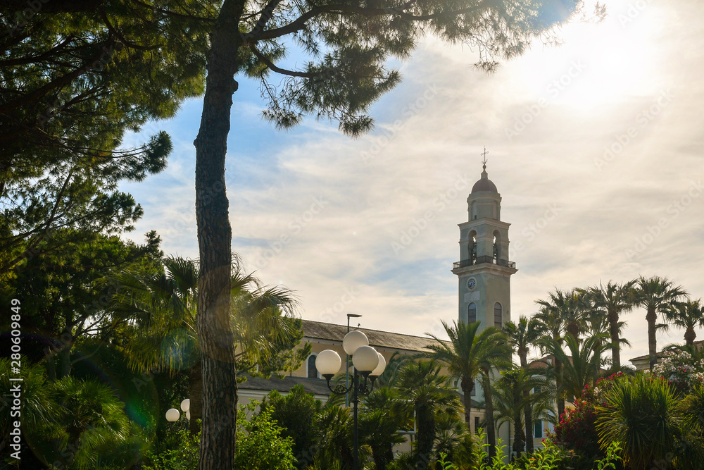 Low angle backlight view of the promenade with pine and palm trees and the bell tower of the parish church of Saint Anthony Abbot (14th century) with lens flare, Diano Marina, Imperia, Liguria, Italy