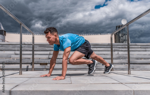 A man is an athlete, abdominal exercises, muscles, summer in city, stretching muscles before jogging, fitness training, sportswear, concentration. Stair background and blue sky.