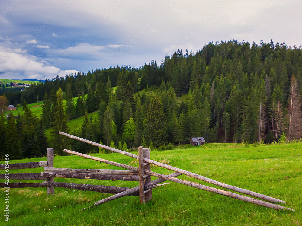 Picturesque spring mountains scene with wooden split rail fence across a green and lush pasture with a old house on the valley surrounded by coniferous woods