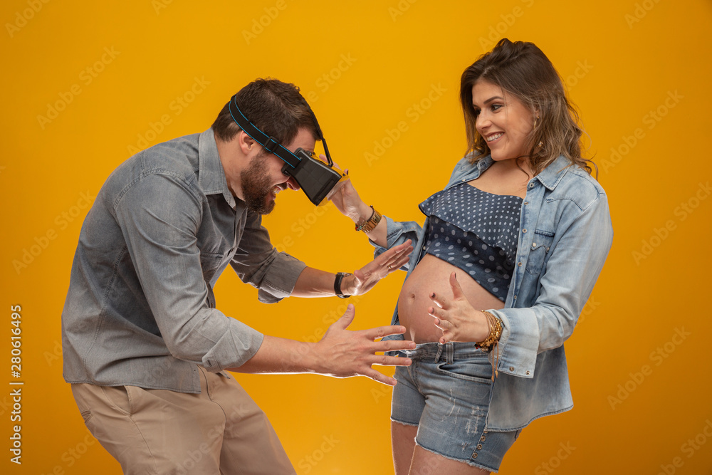 Obraz premium Husband looks at pregnant wife's belly through a VR glasses. Technology simulation to visualize the fetus. Couple joking on yellow background