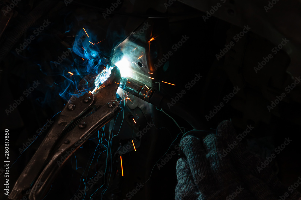 Close-up view strong man is a welder, a metal product is welded with a welding machine in the garage workshop, blue and orange sparks fly to the sides during the body repair car. Auto service industry Close-up view strong man is a welder, a metal product is welded with a welding machine in the garage workshop, blue and orange sparks fly to the sides during the body repair car. Auto service industry