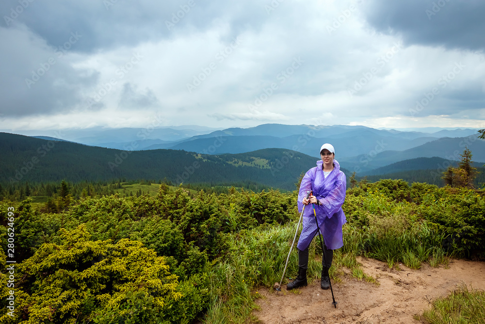 The girl, a tourist with Scandinavian sticks in a raincoat on the background of the beautiful Carpathian mountains. Rise in the mountains. Travel concept, leisure activity, vacation