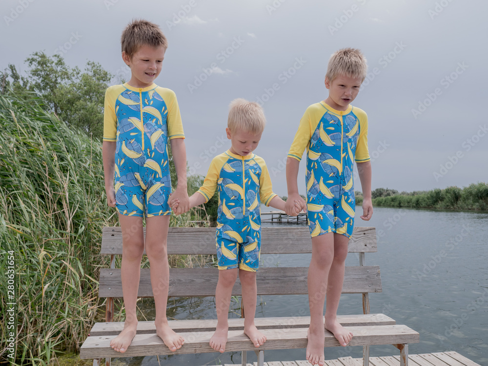 Fototapeta premium Funny kids in bathing suits sit together on wooden bridge near river. boys are happy on vacation in village together. Summer day, river, swimming in the water