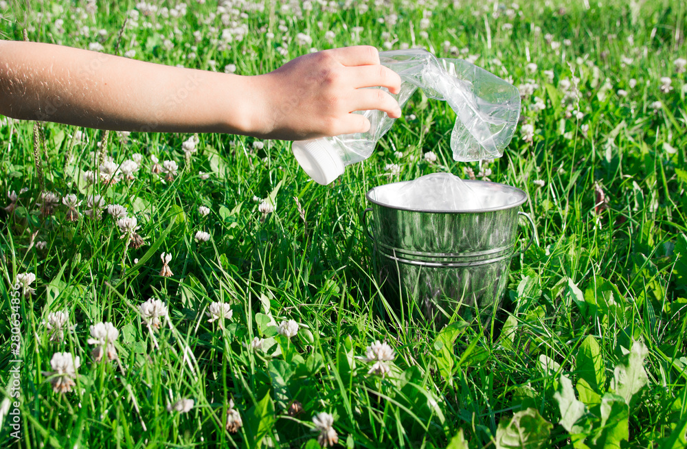 on a green lawn grass with flowers of clover there is a bucket with plastic garbage and a hand collects plastic bottles into it