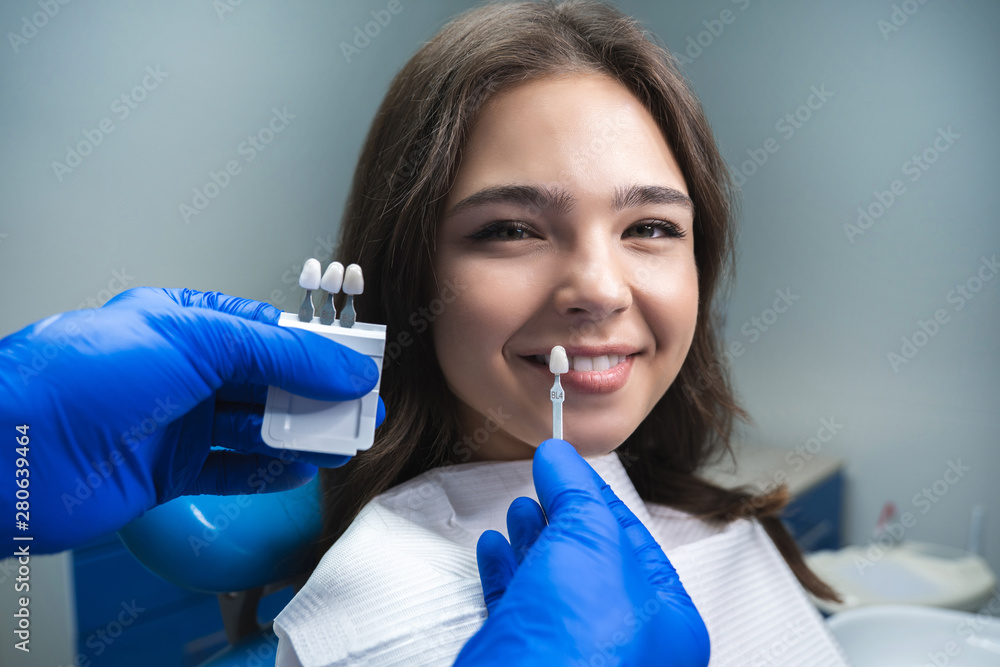 dentist in blue medical gloves applying sample from tooth enamel scale to smiling woman patient to pick up right shade for teeth bleaching procedure dentist in blue medical gloves applying sample from tooth enamel scale to smiling woman patient to pick up right shade for teeth bleaching procedure