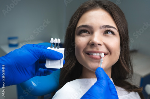 dentist in blue medical gloves applying sample from tooth enamel scale to happy woman patient teeth to pick up right shade for teeth bleaching procedure photo