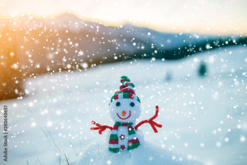 Small funny toy baby snowman in knitted hat and scarf in deep snow outdoor on blurred mountains landscape and falling big snowflakes background. Happy New Year and Merry Christmas greeting card theme.
