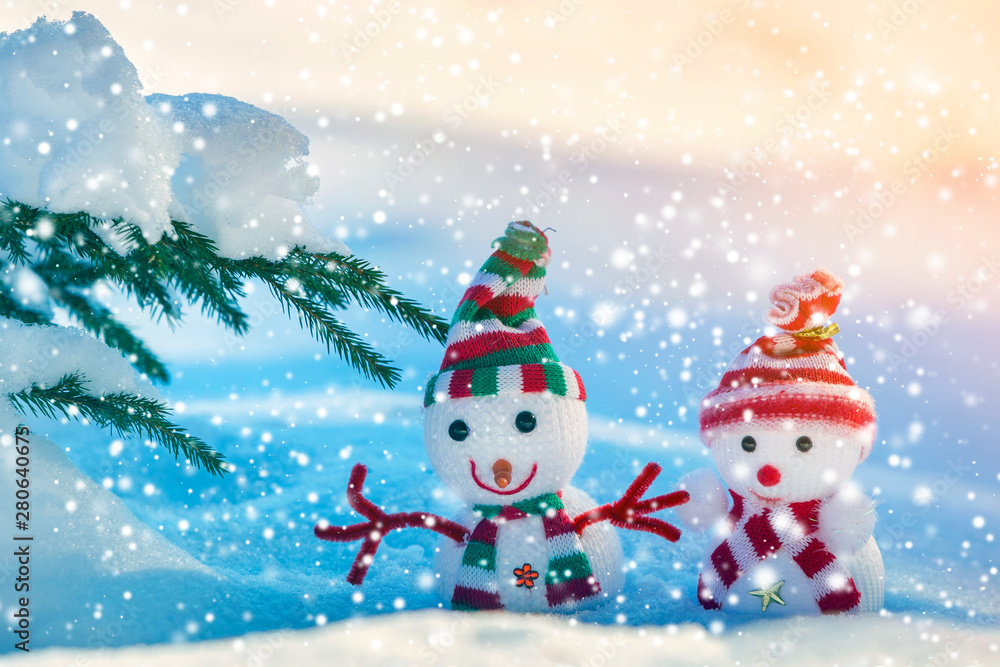 Two small funny toys baby snowman in knitted hats and scarves in deep snow outdoors on bright blue and white copy space background. Happy New Year and Merry Christmas greeting card.