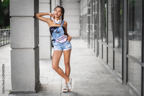 The girl in denim overalls and earpieces around her neck poses near the street wall