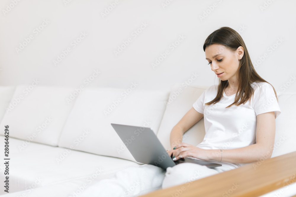Handsome brunette woman using laptop on white couch in light living room Handsome brunette woman using laptop on white couch in light living room