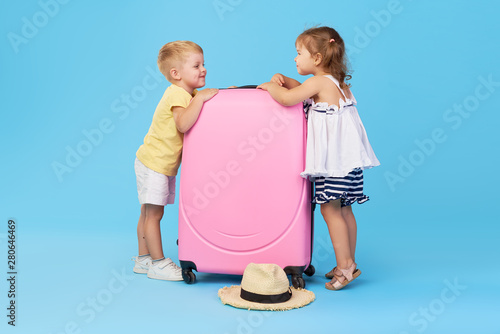 Happy kids holding colorful pink suitcase prepared for summer vacation. Young travelers. Little girl and boy, sister and brother, having fun isolated on blue background