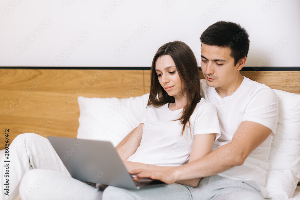 Young tired couple is lying on bed together and looking at laptop