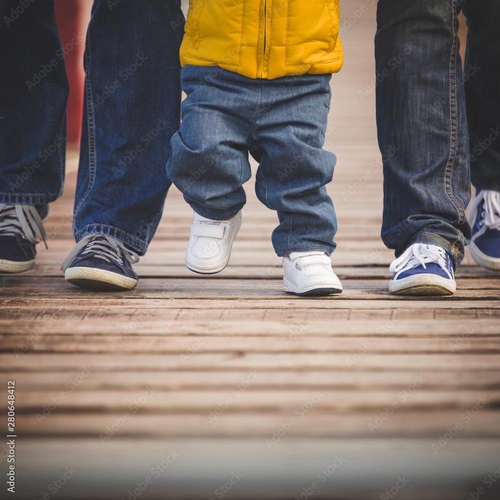 Closeup of legs walking along a wooden pier. Family, mom, dad and little son step on the wooden flooring. People in casual clothes and sneakers. Closeup of legs walking along a wooden pier. Family, mom, dad and little son step on the wooden flooring. People in casual clothes and sneakers.